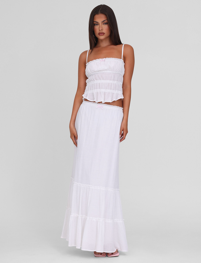A woman with long, straight dark hair and a neutral expression stands against a light gray background wearing the TIGERMIST ASHLYNN TOP - WHITE, featuring adjustable straps and satin bow trim, paired with a long, flowing white skirt.