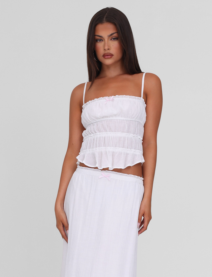 A woman with long dark hair wears the TIGERMIST ASHLYNN TOP in white, featuring adjustable straps and a tiered design, paired with a matching flowy skirt against a plain light gray background.