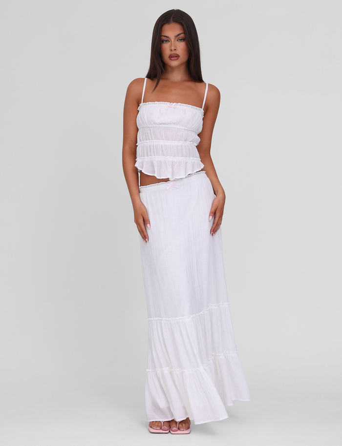 A woman with long dark hair wears the TIGERMIST ASHLYNN TOP in white, featuring adjustable straps and a tiered design, paired with a matching flowy skirt for an effortlessly elegant look against a plain background.