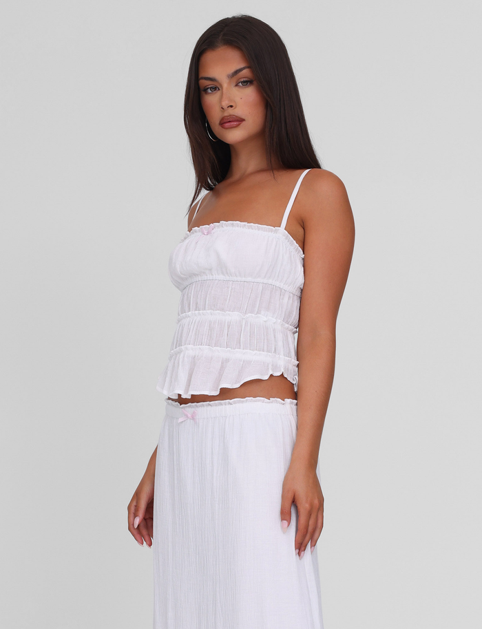 A woman with long dark hair wears the TIGERMIST ASHLYNN TOP in white, featuring adjustable straps and a ruffled hem, paired with a matching skirt, standing against a plain light gray background.