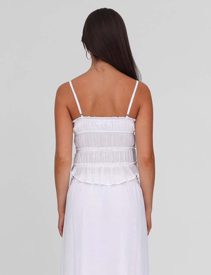 A woman with long brown hair stands facing away, wearing the TIGERMIST ASHLYNN TOP in white, featuring adjustable shoulder straps and a smocked, ruffled bodice. The background is plain light gray.