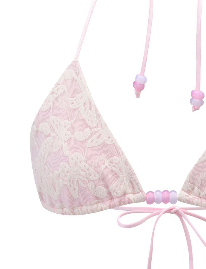 A close-up of the TIGERMIST BRIGITTE BIKINI TOP in ivory lace, featuring floral patterns, pastel beads on adjustable halter straps, and a small pink ribbon bow at the center.