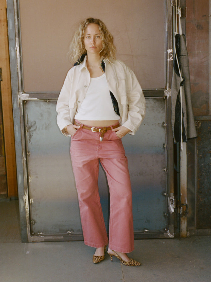 Indoors, a person with a neutral expression stands hands-in-pockets, wearing a white jacket, white tank top, FOUND Canvas Faded Pants with a belt, and leopard print heels.