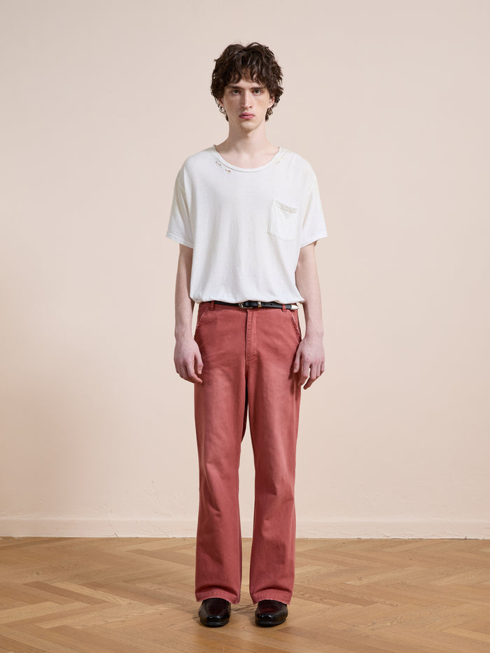 A person stands indoors against a plain beige wall, wearing a white t-shirt, FOUND Canvas Faded Pants in vintage salmon wash, and black shoes on a light wooden floor.