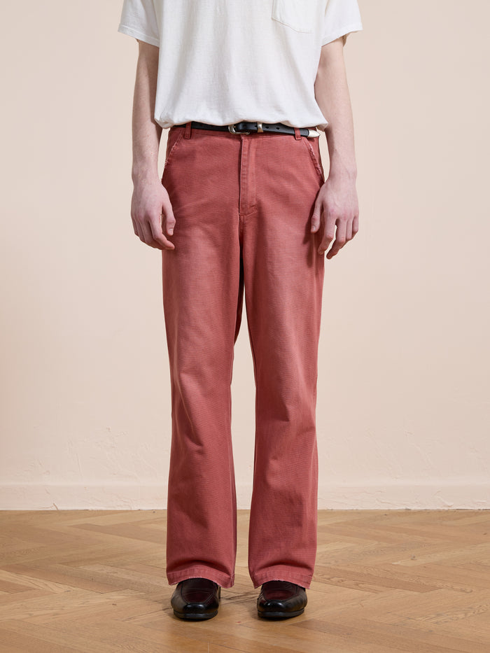 A person stands on a wooden floor against a beige wall, wearing a white T-shirt, FOUND Canvas Faded Pants in vintage salmon wash with a black belt, and dark shoes.